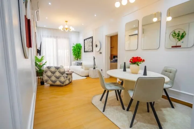 Séjour d'un appartement meublé lumineux : coin salon avec canapé blanc et fauteuil, table ronde et quatre chaises, miroirs au mur, chambre visible au fond.